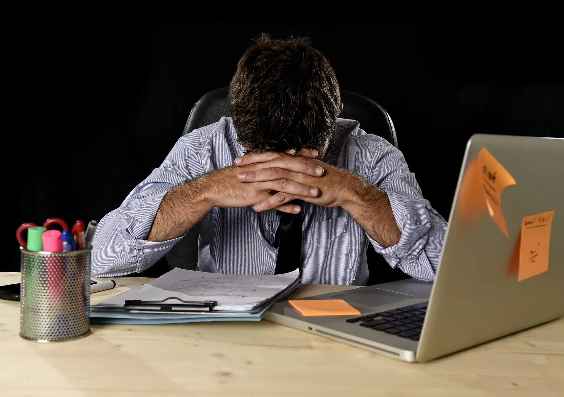 A man sits at a desk with his head in his hands, appearing stressed, surrounded by a laptop, scattered papers, and colorful sticky notes.