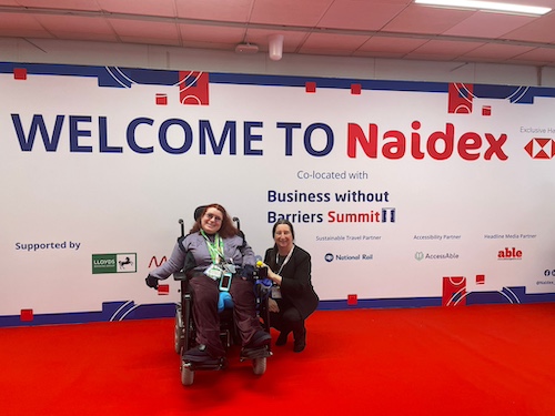 Two people are on a red carpet at the entrance of an event with a "Welcome to Naidex" sign. One person is in a wheelchair, and the other is kneeling beside them, both smiling. Various sponsors' logos are visible in the background.